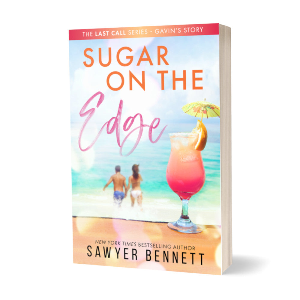 Contemporary romance paperback featuring a couple embracing on the beach in the background, with a hurricane glass with pink liquid and a drink umbrella in the forefront. The title of the book is Sugar on the Edge by New York Times Bestselling Author Sawyer Bennett.