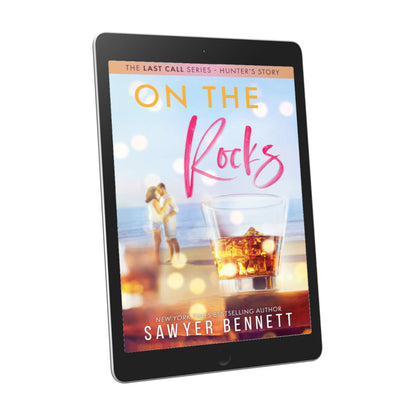 Contemporary romance book cover featuring a couple embracing on the beach in the background, with a highball glass filled with dark liquid and ice in the forefront, displayed on an e-reader. The title of the book is On the Rocks by New York Times Bestselling Author Sawyer Bennett.