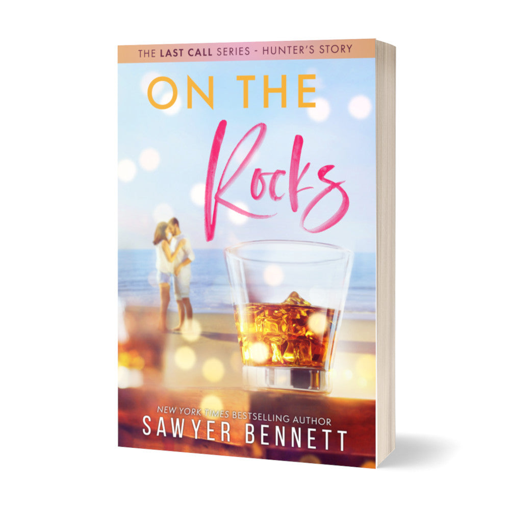 Contemporary romance paperback featuring a couple embracing on the beach in the background, with a highball glass filled with dark liquid and ice in the forefront. The title of the book is On the Rocks by New York Times Bestselling Author Sawyer Bennett.