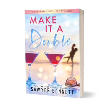 Contemporary romance paperback featuring a couple embracing on the beach in the background, with two martini glasses with pink liquid in the forefront. The title of the book is Make It a Double by New York Times Bestselling Author Sawyer Bennett.