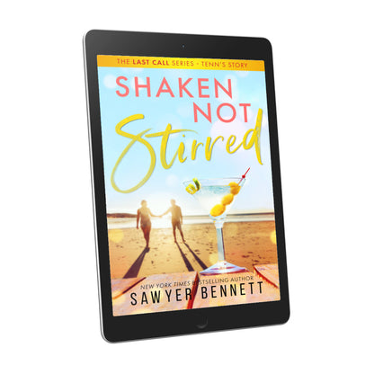 Contemporary romance book cover featuring a couple embracing on the beach in the background, with dirty martini with three olives and a twist of lime in the forefront, displayed on an e-reader. The title of the book is Shaken Not Stirred by New York Times Bestselling Author Sawyer Bennett.