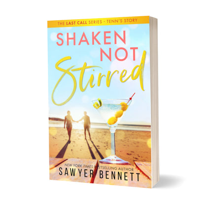 Contemporary romance paperback featuring a couple embracing on the beach in the background, with dirty martini with three olives and a twist of lime in the forefront. The title of the book is Shaken Not Stirred by New York Times Bestselling Author Sawyer Bennett.